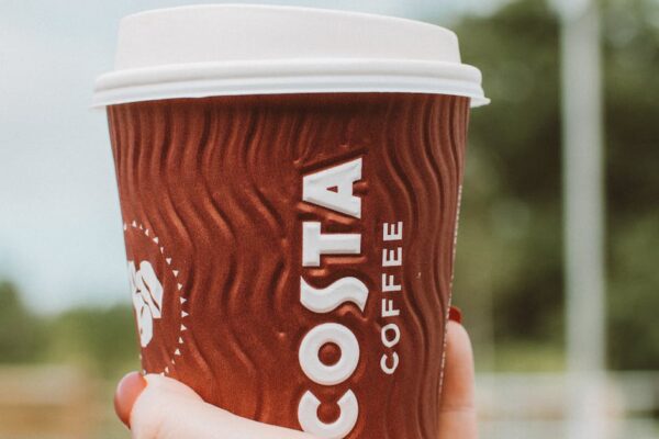 Close-up of a hand holding a Costa Coffee cup in an outdoor setting on a clear day.